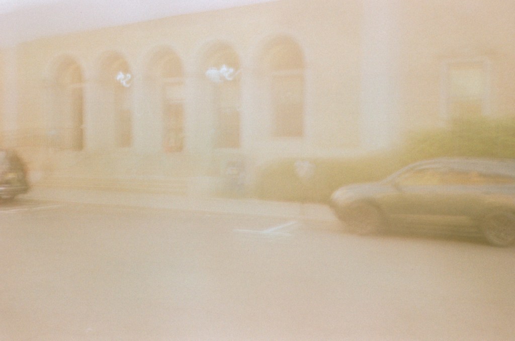 A blurred photograph of a building with arches and a parked car in the foreground, suggesting motion due to the lack of sharpness.